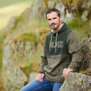 Man sitting on a rock in front of rocky hill wearing blue jeans and a Ridgeline South Island Hoodie in a two-tone Olive colour