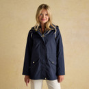 A woman wearing a navy waterproof hooded jacket with gold poppers and front flap pockets, styled with light trousers against a neutral background.