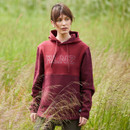 Woman standing in long grass, in brown leggings and a Ridgeline South Island Hoodie in a two-tone Winterberry Red colour