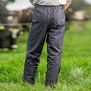 Person wearing dark rain trousers stands in a grassy field with hands in pockets, cows grazing in the blurred background, conveying a rural, outdoorsy feel.