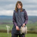 Woman in a blue coat holds a spool of wire outdoors with rolling green hills in the background, conveying a sense of rural work and resilience.