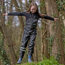 A girl balances with arms outstretched on a grassy slope in a forest. She wears olive bib overtrousers and blue boots, conveying focus and adventure.