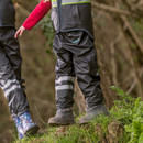 Two children in outdoor gear and rain boots stand on a grassy edge in a wooded area. One wears a red sleeve; both have waterproof pants and boots.