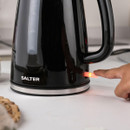 A person's finger presses the power button on a black Salter electric kettle.