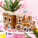 Gingerbread train with white icing detail on round platter, surrounded by red-striped candies. Festive atmosphere with blurred pink tree and greenery.