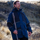 A man in a blue jacket stands outside on a grassy hillside, holding a wooden stick. The lighting is soft, suggesting a calm and serene outdoor setting.
