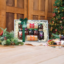 A festive wooden table holds a gourmet gift box with preserves and oatcakes, adorned with a green bow. A decorated Christmas tree glows softly nearby.