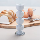 A stack of light blue egg cups on a table, with sliced bread and eggs in the background.