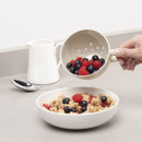 Hand pouring mixed berries from a colander into a bowl of cereal on a counter with a spoon and jug nearby.