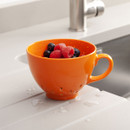 An orange colander cup filled with fresh raspberries and blueberries on a wet counter.