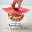 A finger pressing the centre of a red silicone lid on a bowl of salad, indicating an airtight seal.