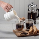 Pouring milk into coffee from a white porcelain churn jug, with a French press and biscotti on a wooden tray.
