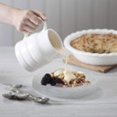 A hand pouring custard from a white porcelain churn jug over a crumble dessert with blackberries on a glass plate, pie in background.