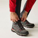 Close-up of grey hiking boots being fastened, featuring red laces, supportive ankle collars and rugged black and red grip soles.