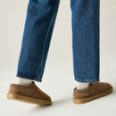Rear view of taupe suede slip-on shoes worn with white socks and straight-leg blue jeans, showing the low-profile design and tan soles.