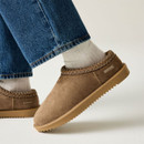 Close-up of a taupe suede slip-on shoe with decorative braided trim, soft upper and ridged tan sole.