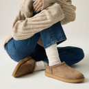 Taupe suede slip-on shoes styled with white socks, blue jeans and a cream knitted jumper in a relaxed seated pose.