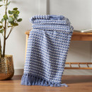 A blue and white stab stitch design throw with frilled ends folded and raped over a wooden stool