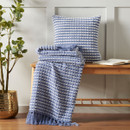 A blue and white stab stitch design throw with frilled ends with a matching cushion folded and raped over a wooden stool