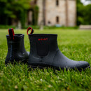 Navy Ariat ankle rain boots with red detailing sit on lush green grass. The boots have water droplets on them, with a blurred building and trees in the background. Navy Ariat ankle rain boots with red detailing sit on lush green grass. The boots have water droplets on them, with a blurred building and trees in the background.
