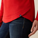 A person wearing a bright red sweatshirt and dark blue jeans, with a focus on the sweatshirt's hem and a small logo tag. The scene conveys casual comfort. A person wearing a bright red sweatshirt and dark blue jeans, with a focus on the sweatshirt's hem and a small logo tag. The scene conveys casual comfort.