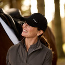 Woman in a black "Ariat" cap smiles toward a horse, wearing a grey jacket. Background features blurred trees, creating a warm outdoor atmosphere.