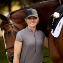 A smiling person wearing a black cap and grey shirt stands beside a brown horse. The setting is outdoors with a warm, serene atmosphere.