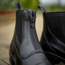 Close-up of sleek black leather boots with a front zipper detail, set on a wooden surface. The background is blurred, highlighting the boots' polished texture.