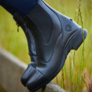 Close-up of sleek, black leather ankle boots on a wooden fence. The background shows tall green grass, conveying a sense of outdoor adventure.