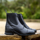 A pair of sleek black leather ankle boots with zippers and pull tabs sits on a wooden surface. The background shows a blurred green foliage, conveying an outdoor feel.