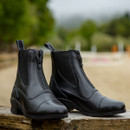 Black leather equestrian boots with front zippers rest on a wooden surface. Blurred green and sandy background suggests an outdoor setting.
