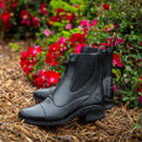 Black leather ankle boots with zippers on wood chips, surrounded by vibrant red and pink flowers. The scene conveys elegance and nature.