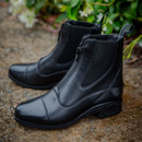 Black leather ankle boots with front zippers, sitting on a textured ground. Lush green foliage in the background adds a natural, earthy feel.