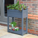 A grey wooden trough with herbs growing in the deep tray at the top and a shallow tray at the bottom, positioned against an outdoor wall.