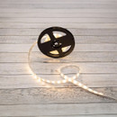 A glowing LED strip light unspools from a black reel, casting a warm, soft light on a light wooden surface, creating a cosy and inviting atmosphere.