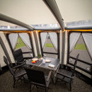 Inside a cosy tent with large windows, a table is set with plates, fruit, and drinks. Four black camping chairs surround the table, creating a warm, inviting atmosphere.