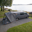 Van with attached tent parked by a lakeside with trees and grassy area.