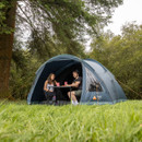 A couple is seated at a table inside a blue dome tent, enjoying drinks. The tent is set on lush green grass under tall trees, conveying a serene camping atmosphere.