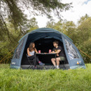 A man and woman sit smiling at a table inside a large blue tent, surrounded by lush green grass and trees, conveying a relaxed camping vibe.