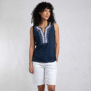 A person stands confidently, wearing a navy sleeveless top with white embroidery and white shorts against a simple grey background.