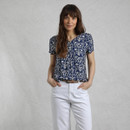 A person stands confidently, wearing a navy patterned top and white pants, with hands casually placed in pockets against a textured background.