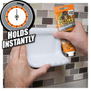 Hands applying adhesive from a Gorilla Glue tube to mount a shelf on tiles. Text: "Holds Instantly".
