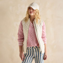 A woman modelling a cream quilted waistcoat layered over a pink V-neck cable-knit jumper and blue-and-white striped trousers.