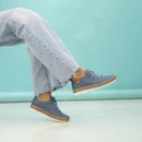 A person wearing blue shoes and frayed jeans with crossed ankles, suspended in air against a teal background.