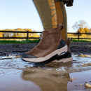 Person in brown boots stepping into a puddle, with autumn trees in the background.