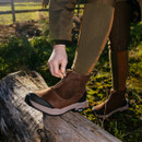 Person tying a brown boot outdoors on a log with greenery in the background.