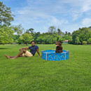 A view of a brown dog in a blue dog paddling pool with a man laid down next to it on green grass