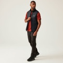 A full length view of a man wearing a black gilet over a red t-shirt with brown trousers and walking boots.