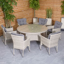 Outdoor patio set with a round wicker table and six chairs featuring gray cushions. Set against a wooden fence, with potted plants in the background.