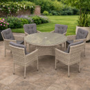Outdoor patio set with a round glass-top table and six wicker chairs with gray cushions. Surrounded by lush green garden and flowering plants.